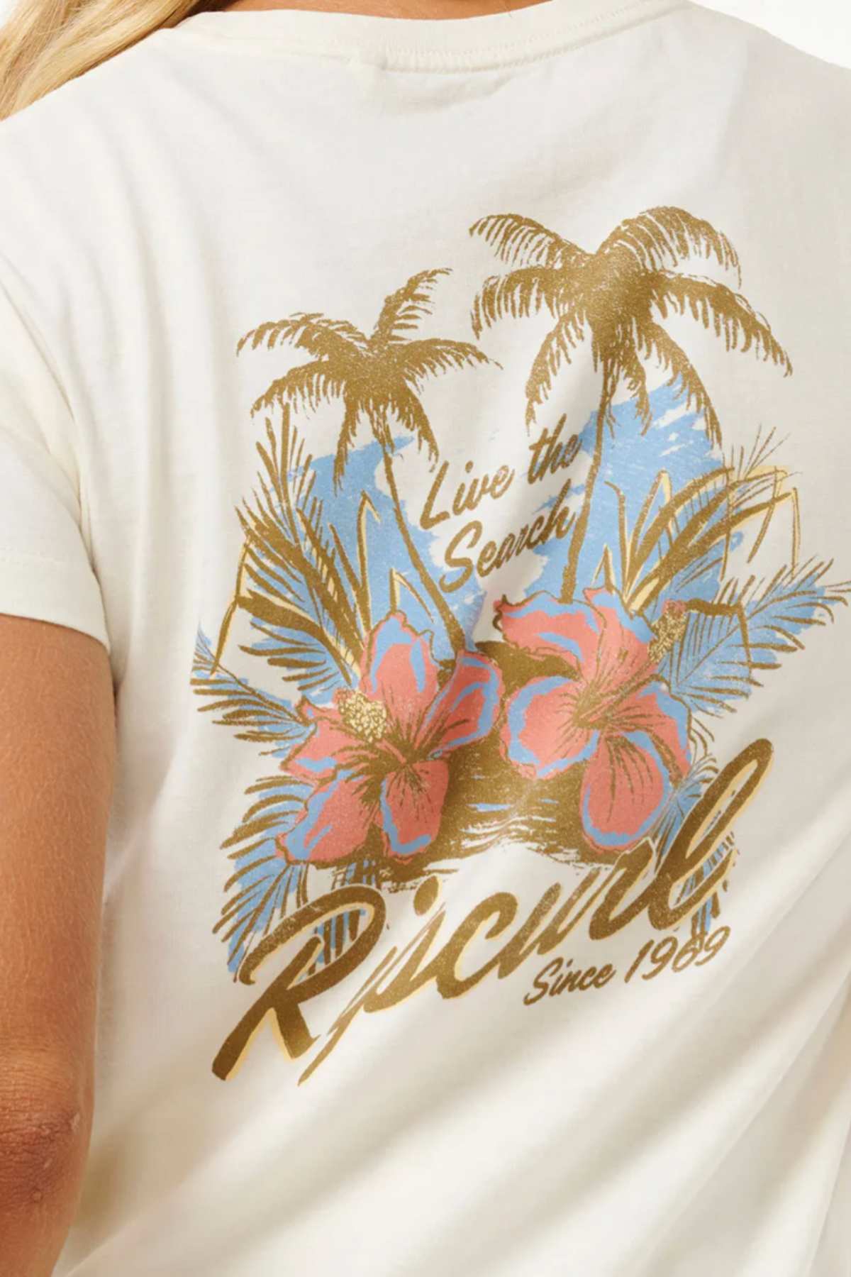 The image showcases the back of a white Rip Curl Wild Bloom Standard Tee, ideal for adding to your casual wardrobe. Made entirely from 100% cotton, it features two large pink hibiscus flowers, palm trees, and a backdrop of a blue sky. The design includes the text "Live the Search" at the top and "Rip Curl Since 1969" at the bottom.