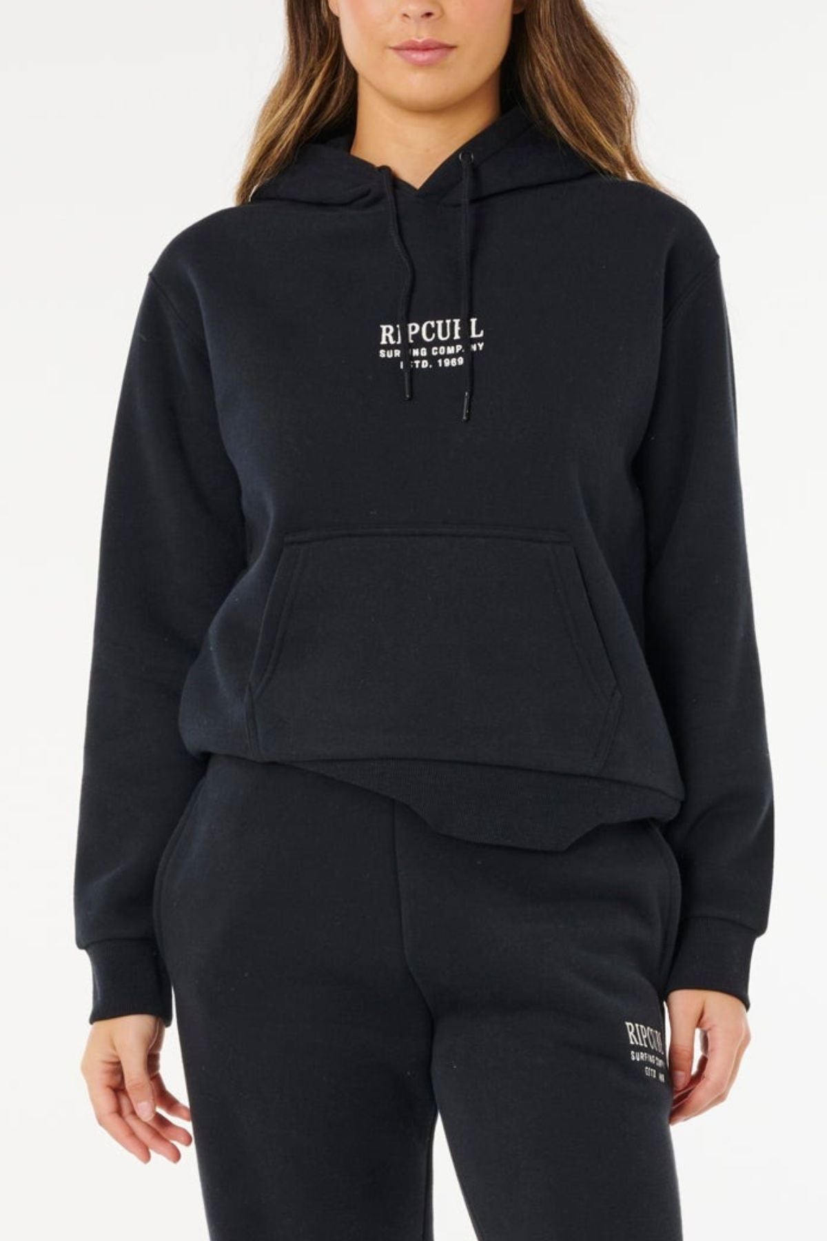 A woman stands against a plain white background wearing the Rip Curl Surf Staple Relaxed Hood and matching black sweatpants, both with small white logosβshowcasing classic surf heritage style.