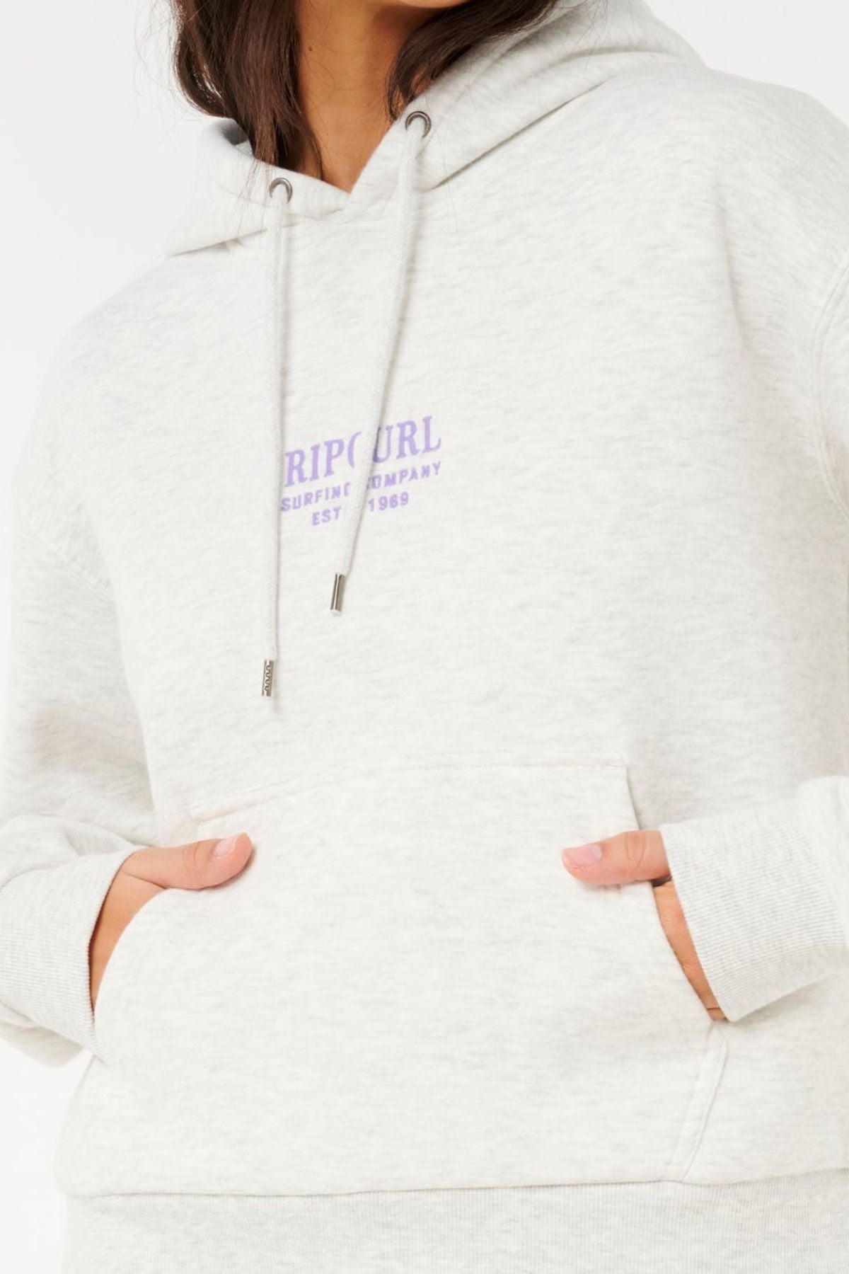 A person in a Rip Curl Surf Staple Relaxed Hood with "Rip Curl Surfing Company Est. 1969" in purple on the chest, hands in the front pocket. The classic surf heritage look is shown without revealing their face.