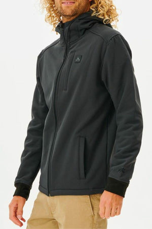A person with long, curly blonde hair wears the black Rip Curl Anti Series Soft Tech Fleece jacket with a hood and beige pants. The water-resistant jacket has zippered pockets, a small chest logo, and is shown against a plain white background.