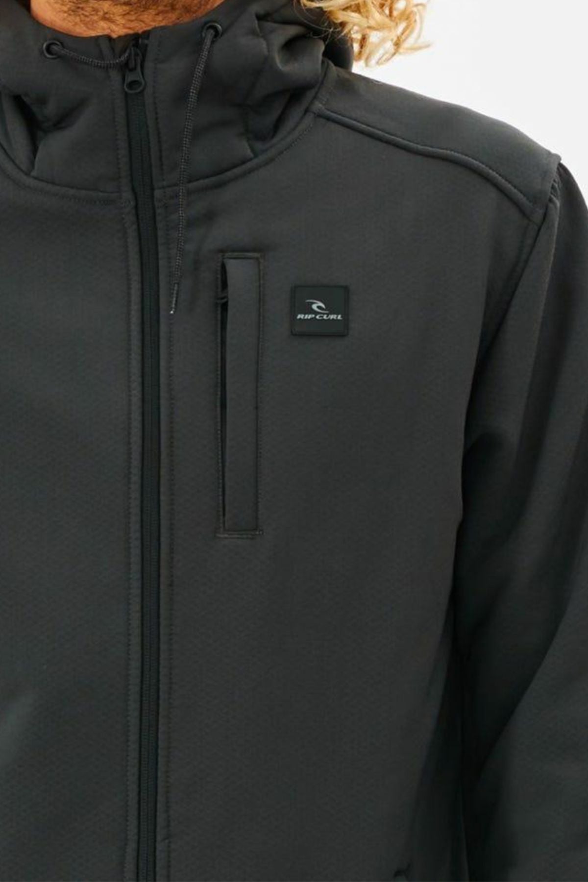 Close-up of a person wearing a black Rip Curl Anti Series Soft Tech Fleece jacket with hood and zippered chest pocket. The face is hidden, with blond hair visible on the right side.