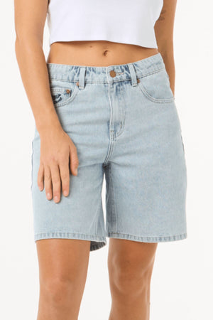 Model wearing Rip Curl Aggie Relaxed Denim Short in light blue and a white cropped tank top, standing against a plain white background. Only the torso and upper legs are visible.