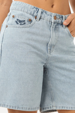 A person wearing Rip Curl Aggie Relaxed Denim Shorts with one hand in a pocket. The light blue shorts have a relaxed fit, button and zipper closure, belt loops, and an embroidered design above the front pocket.