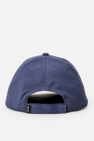 The Rip Curl Surf Staple Cap is a navy blue, six-panel baseball cap with an adjustable strap and a small black tag on the left side of the back opening, shown against a plain light gray background.