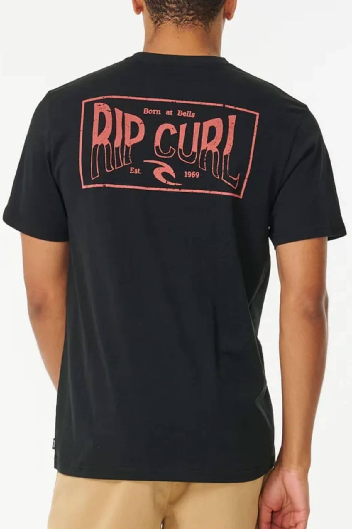 Wearing a black 100% cotton Rip Curl Affinity Tee featuring a bold red "RIP CURL" logo, "Born at Bells," "Est. 1969," and wave graphic on the back, paired with beige shorts, the person stands facing away from the camera.
