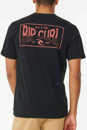 Wearing a black 100% cotton Rip Curl Affinity Tee featuring a bold red "RIP CURL" logo, "Born at Bells," "Est. 1969," and wave graphic on the back, paired with beige shorts, the person stands facing away from the camera.