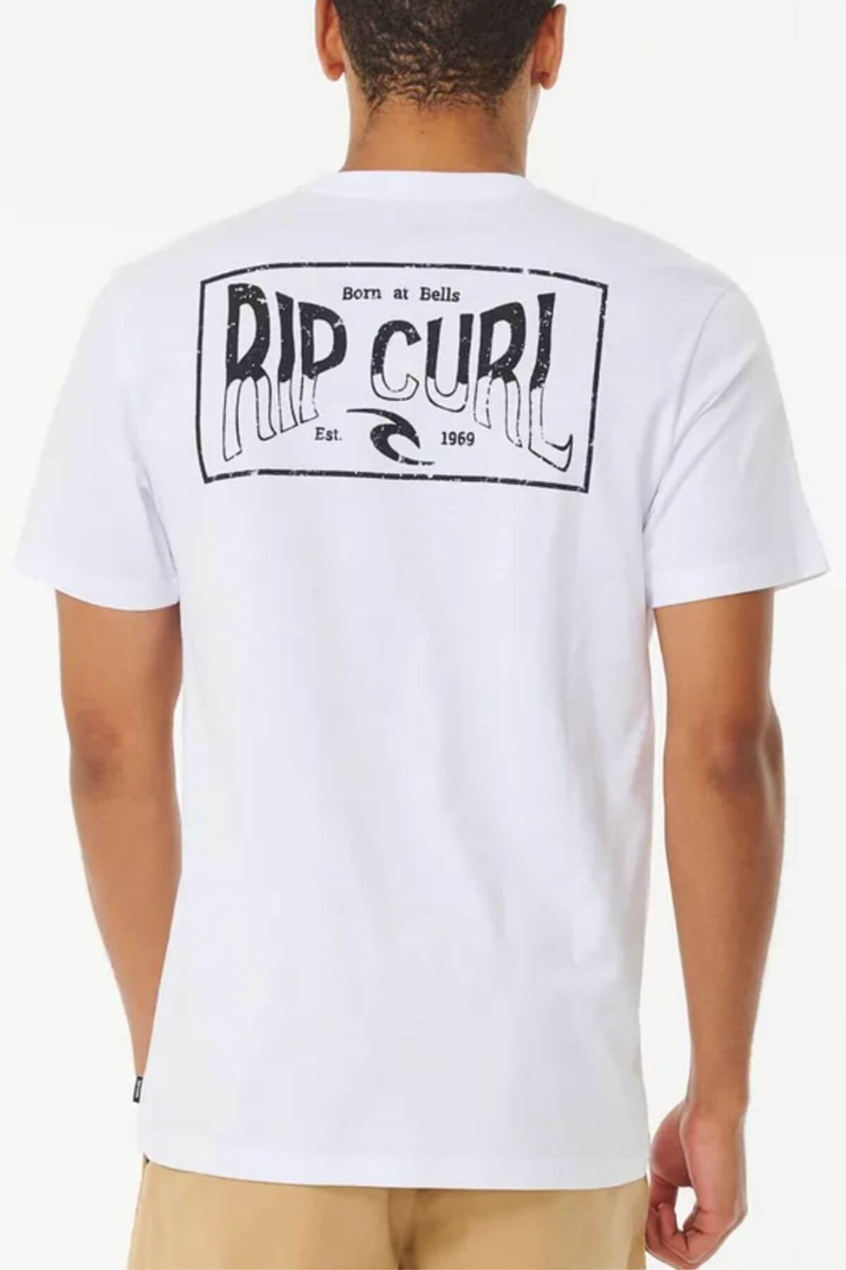 The image displays a person wearing the Rip Curl Affinity Tee, featuring a large "RIP CURL" logo with "Born at Bells Est. 1969" on the back. This white, 100% cotton tee pairs with beige pants for a casual look and provides a standard fit.