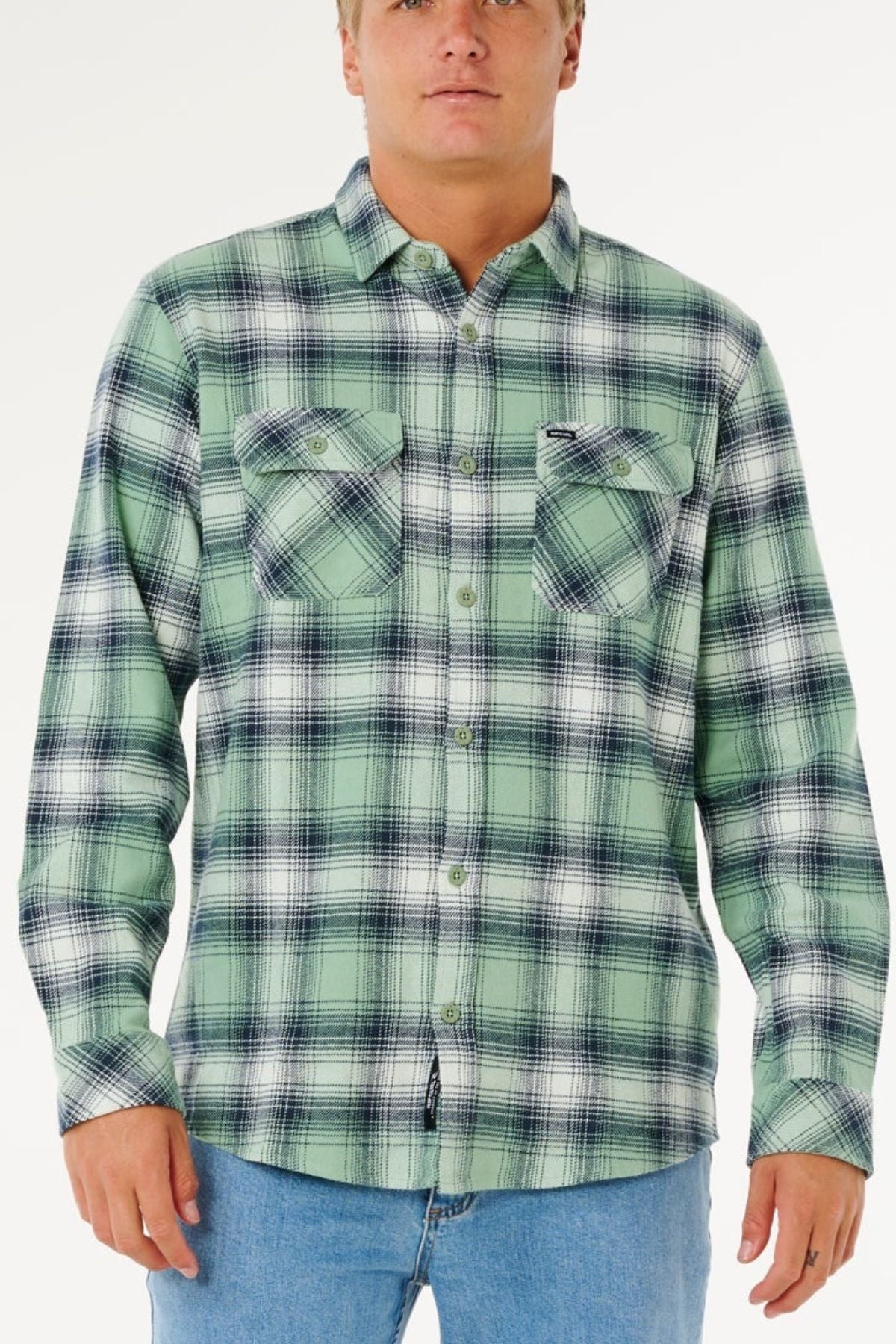 A person wears the Rip Curl Count Flannel Shirt in light green and black plaid with two chest pockets, paired with light blue jeans, standing against a plain white background.