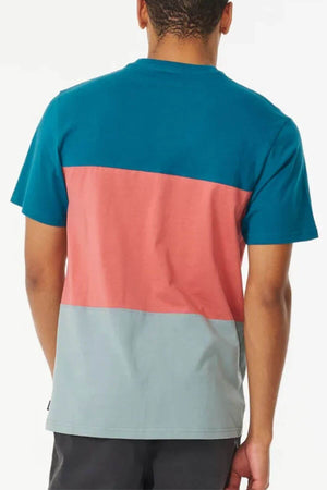 A person stands with their back to the camera, wearing the Rip Curl Divided Tee—a crew neck shirt with three horizontal color blocks: teal, pink, and light gray.