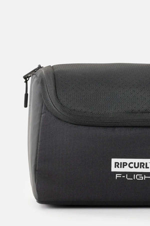 A close-up of a black Rip Curl F-Light Toiletry bag on a white background showcases its multiple compartments, zipper closure, ventilated mesh section, and prominently printed side logo.