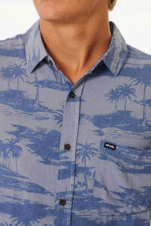 A person wears a Rip Curl Party Pack Short Sleeve Shirt in blue, featuring a tropical palm tree and island pattern, collar, chest pocket with black and white label; their face is partially hidden.