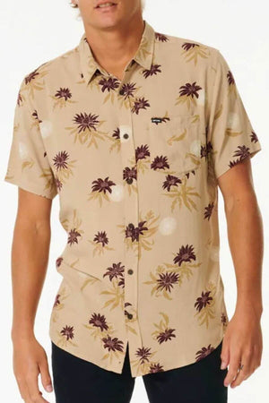 A person wears a beige Rip Curl Sun Razed Floral Short Sleeve Shirt, featuring purple and yellow floral designs and a single chest pocket, paired with black pants against a plain white background.