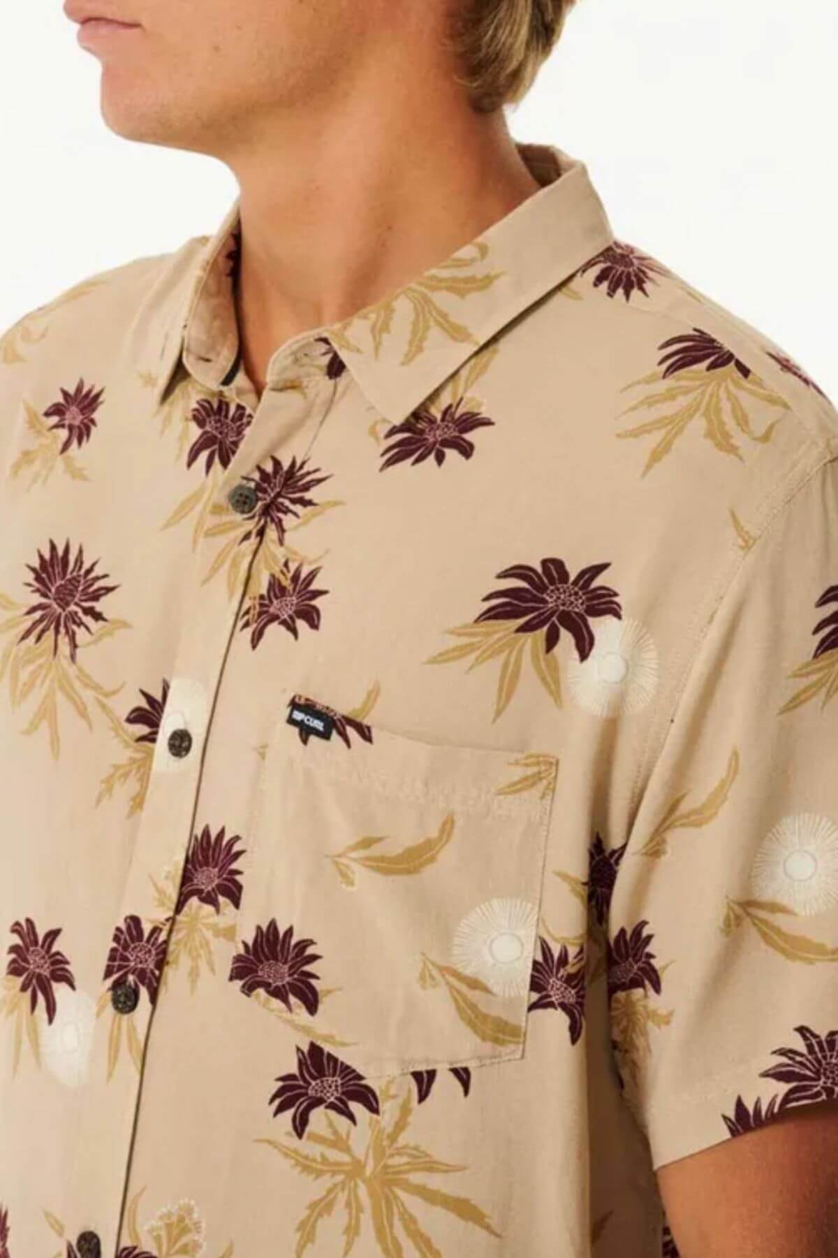 Someone is wearing a Rip Curl Sun Razed Floral Short Sleeve Shirt in beige with maroon and golden floral patterns, featuring a front pocket with a small black label. The background is plain white.