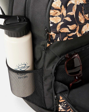 A close-up of the Rip Curl Chaser 33L Backpack with a floral leaf pattern, featuring a white water bottle in the mesh side pocket and brown sunglasses on the zipper. This backpack fits a 15-inch laptop.