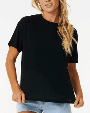 A person with long blonde hair is wearing a plain black Rip Curl Classic Relaxed Tee and light blue denim shorts. They are standing against a white background, holding the bottom hem of the shirt made of soft cotton jersey with their left hand, and appear to be smiling slightly.