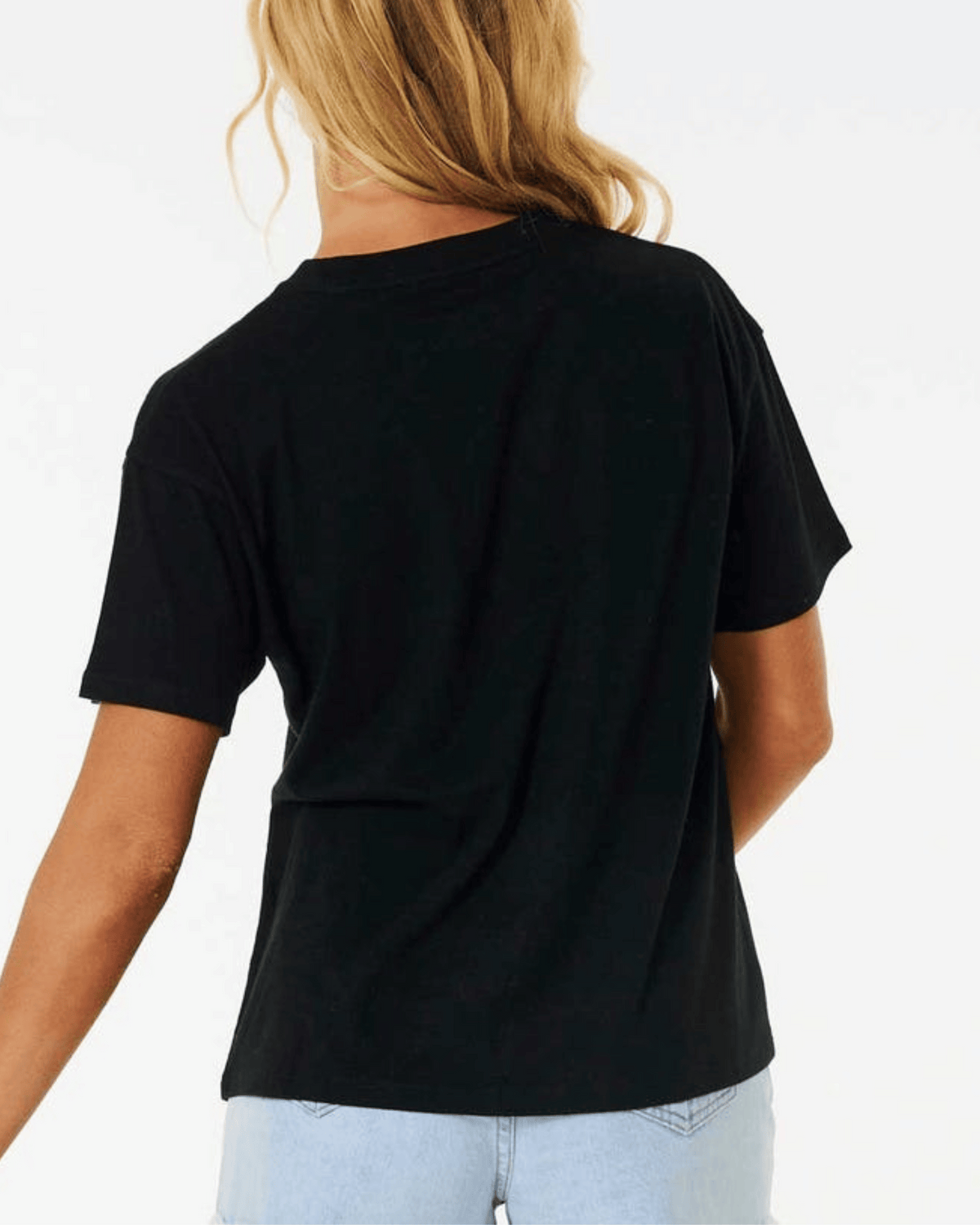 A person with long, blonde hair is shown from the back, wearing a plain black Rip Curl Classic Relaxed Tee and light blue jeans. The comfortable cotton jersey of the tee makes it perfect for everyday wear. The background is white.