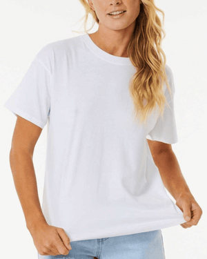 A person with long, wavy hair is wearing a Rip Curl Classic Relaxed Tee in Optical White paired with light blue jeans, exuding a casual and laid-back vibe. They are standing against a white background, gently holding the side of their cotton jersey T-shirt.