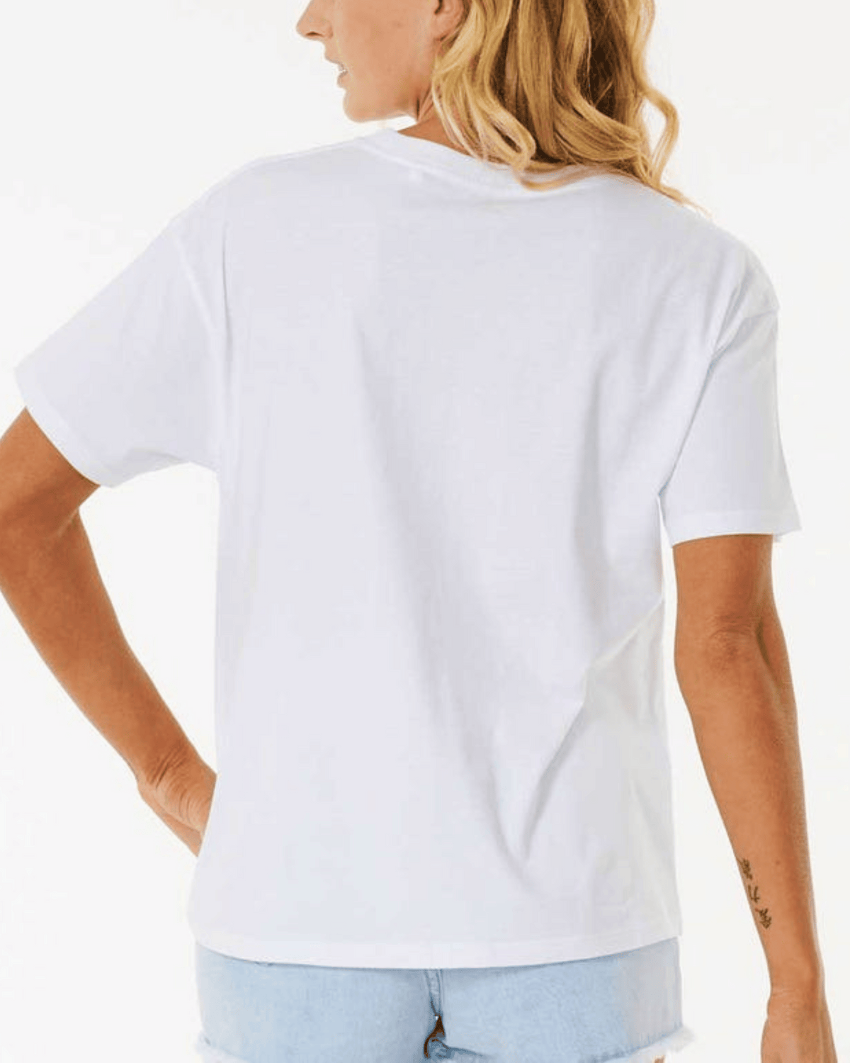 A person with long blonde hair, dressed in a casual and laid-back white Rip Curl Classic Relaxed Tee paired with light blue denim shorts, is seen standing with their back to the camera. Their left hand is resting on their hip, showcasing a tattoo on their right forearm.