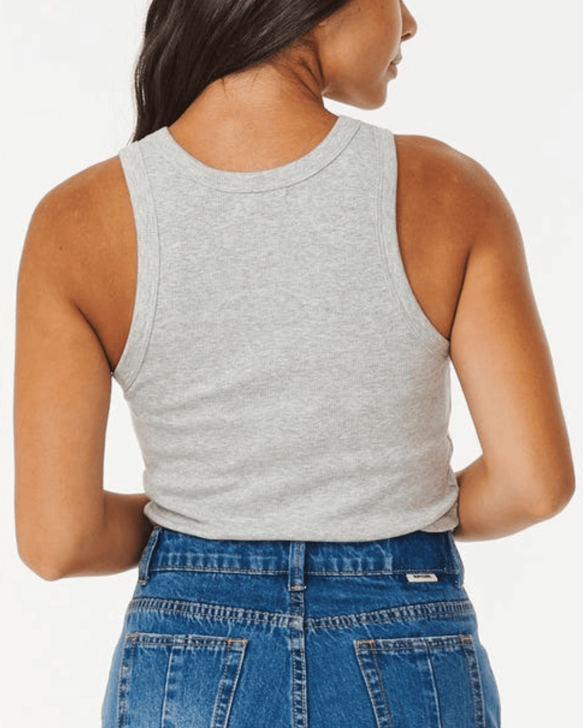 A person with long hair, facing away from the camera, wears the Rip Curl Classic Ribbed Tank in gray paired with blue jeans. The cotton elastane blend offers comfort and style against a plain white backdrop.