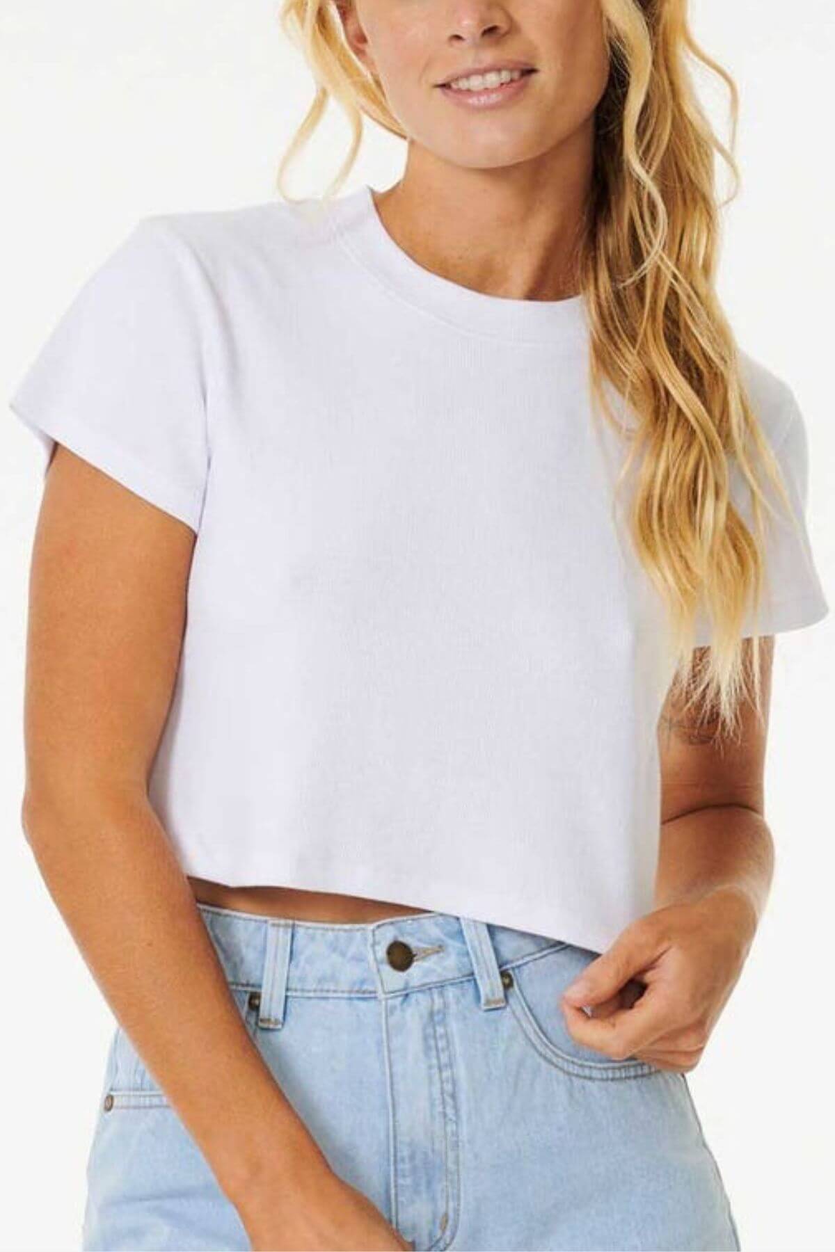 A woman in a Rip Curl Classic Ribbed Tee and light blue jeans smiles, her long, wavy blonde hair cascading against a plain, light background that highlights this wardrobe essential's timeless classic style.