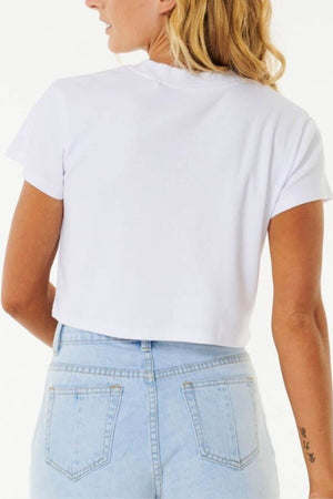 A person with blonde hair, viewed from behind, wears a Rip Curl Classic Ribbed Tee in white and light blue high-waisted jeans. A small tattoo on their left arm adds intrigue, while the plain white background highlights this wardrobe staple.
