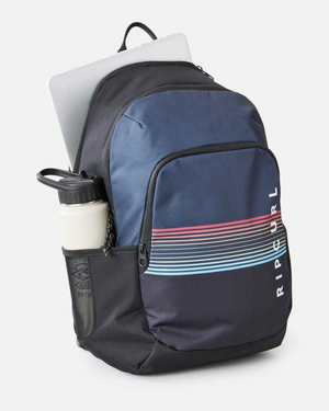 The Rip Curl Ozone 30L School Backpack in blue and black, made from recycled polyester, showcases colorful stripes. It includes a front pocket, a side mesh pocket with a water bottle, and has space for a laptop in the main compartment, making it perfect for school.
