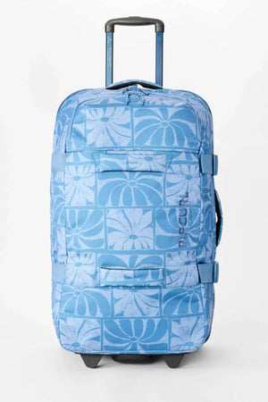 The Rip Curl F-Light Artist Of The Search Ty Williams 100L Global Travel Bag is a blue rolling travel bag with a telescopic handle, wheels, side straps, front zip pocket, and a lighter blue geometric floral pattern.