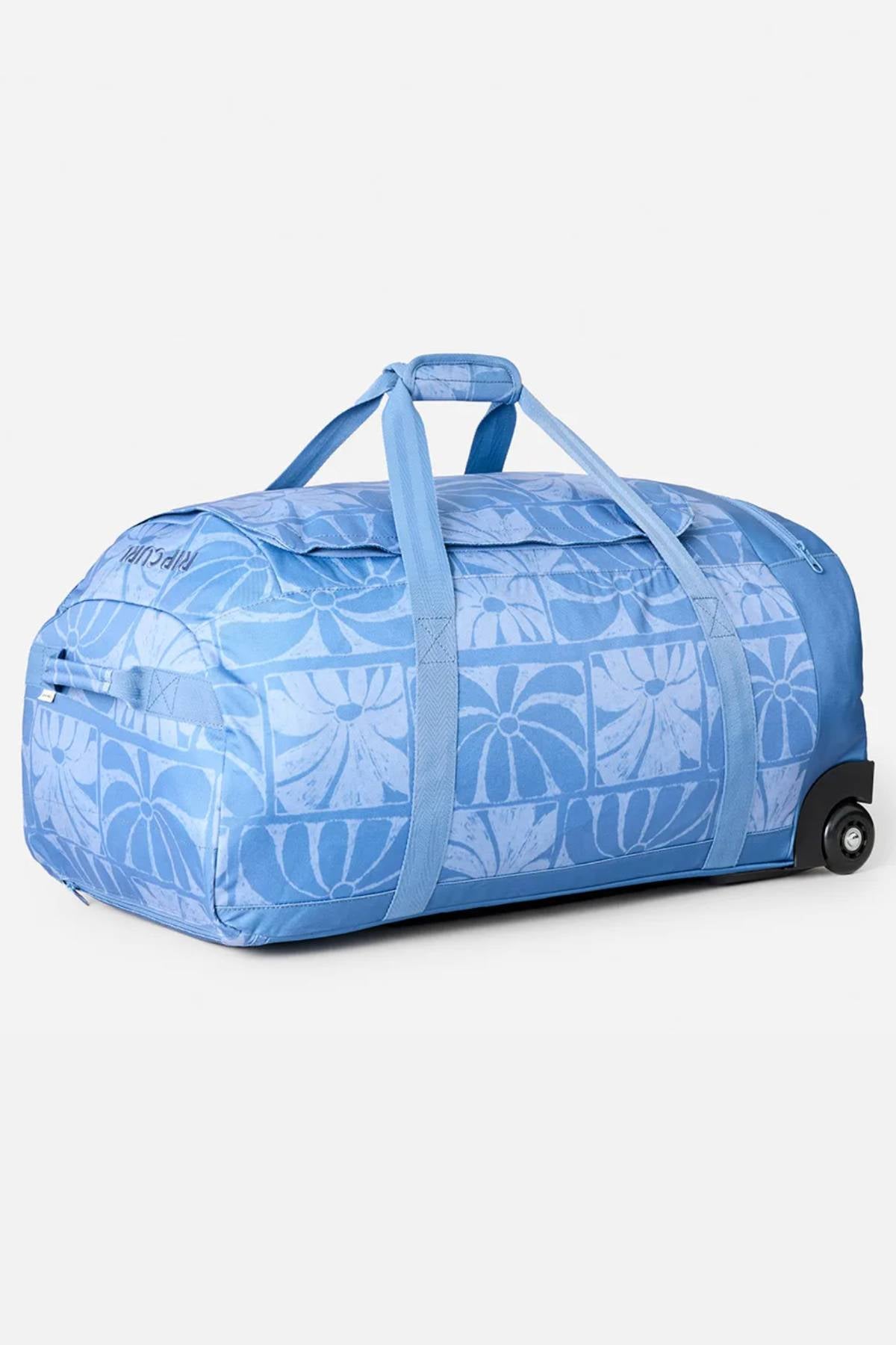 The Rip Curl Jupiter 80L Mixed Travel Bag is a large, light blue travel bag with a patterned design, two handles, a zippered top, wheels for easy rolling, and is made from recycled polyester. Shown against a plain white background.