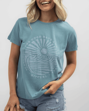 A smiling person wears the Rip Curl Good Waves Standard Tee made of cotton jersey, with a circular wave and setting sun design. Text reads "Searching for Good Days and Good Waves, Ripcurl." They pair it with distressed denim shorts.