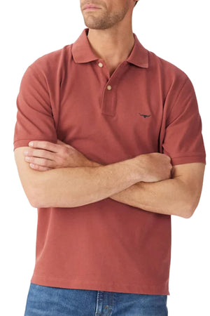 A man stands with arms crossed, wearing the rust-colored RM Williams Rod Polo featuring a Longhorn chest embroidery, paired with blue jeans. His face is partially out of frame.