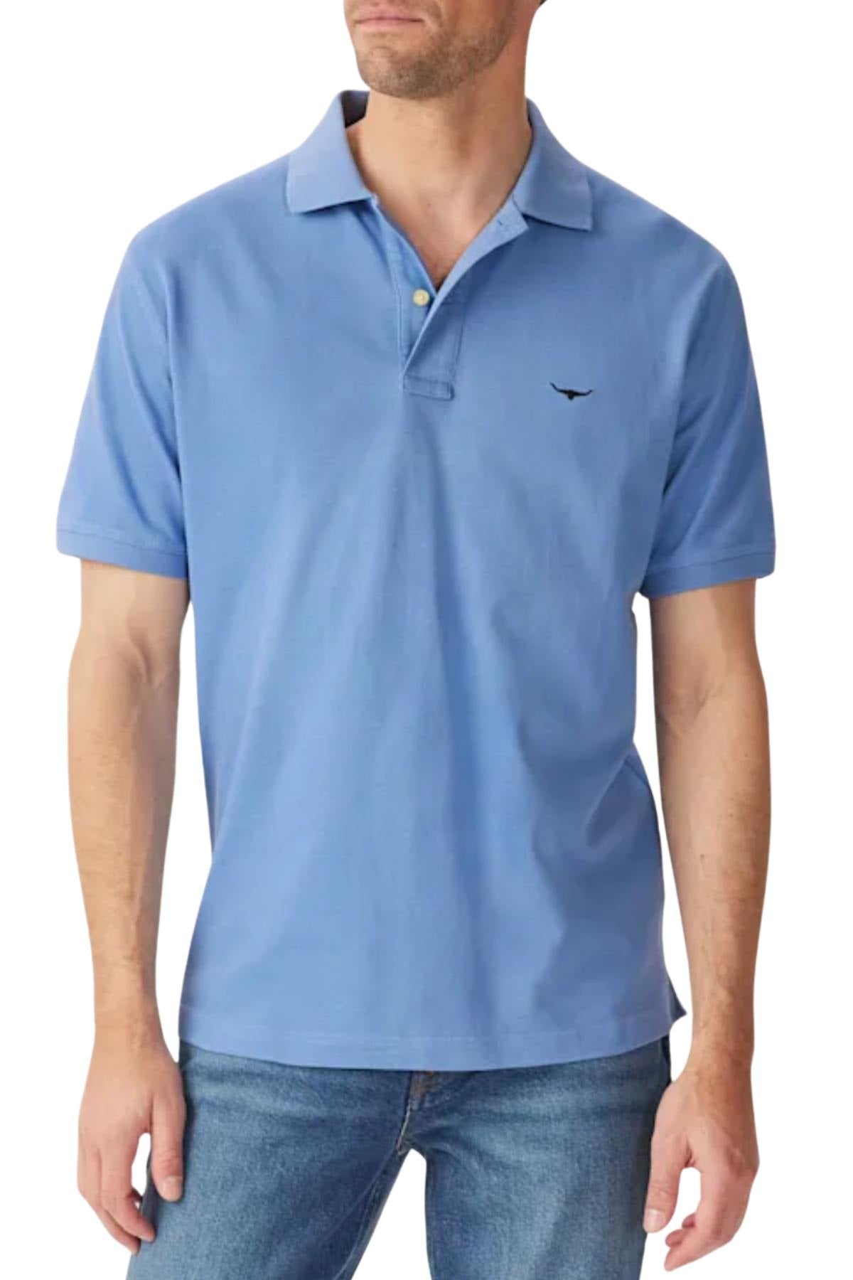 A man wears the RM Williams Rod Polo in light blue with a small black logo on the left chest, paired with blue jeans. His face is partially out of frame as he stands against a plain white background.