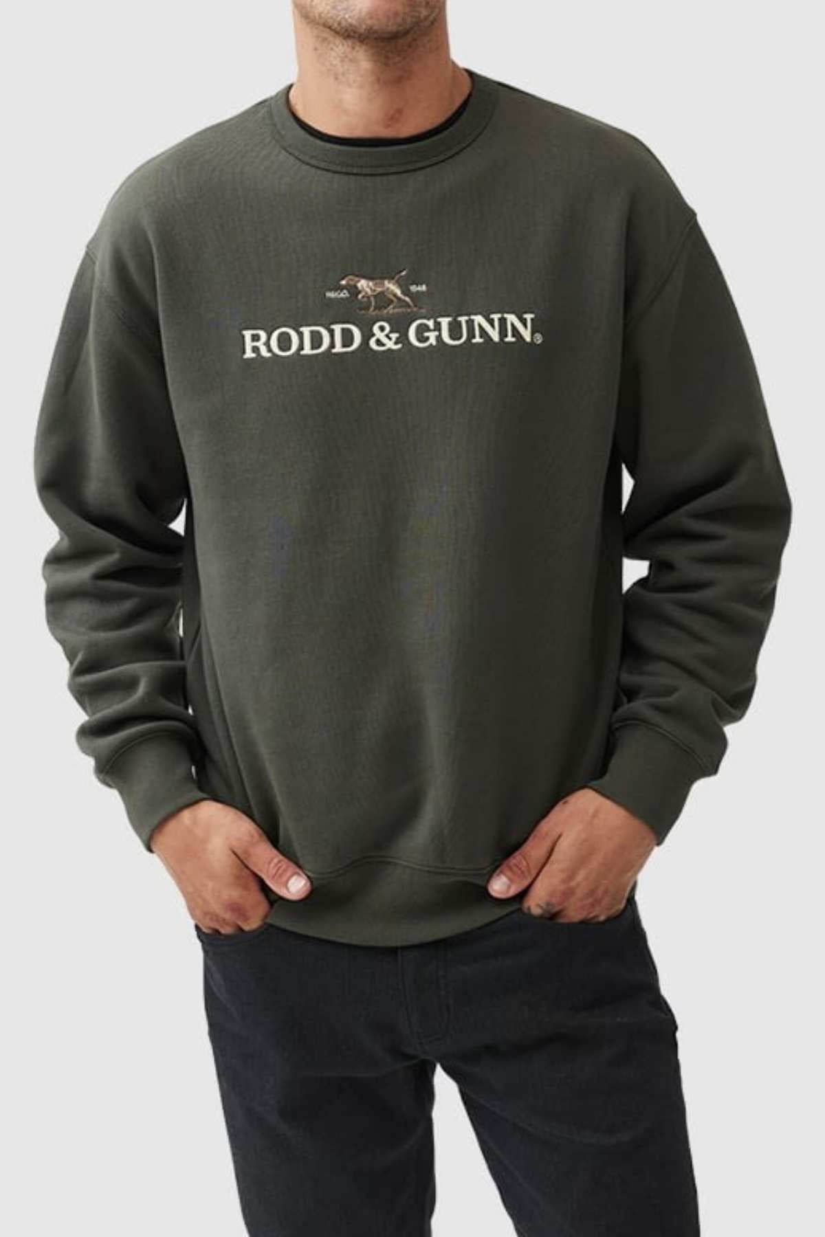 A man wears a Rodd & Gunn Gunn Logo Sweat crafted from premium USA cotton in dark green, showcasing the iconic dog emblem on the front. He pairs it stylishly with dark pants and casually slips his hands into his pockets.