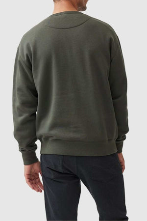 A person stands with their back to the camera, wearing a dark green Rodd & Gunn Gunn Logo Sweat and black pants. The background is a plain light color.