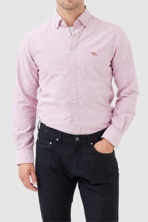 A man wears the Rodd & Gunn Gunn Oxford Sports Fit Shirt, a stylish 100% cotton light pink long-sleeve button-up, paired with black pants, standing with one hand in his pocket against a plain light gray background.