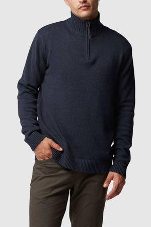 A person is dressed in a Rodd & Gunn Merrick Bay Knit sweater in dark navy, featuring long sleeves and a premium YKK half-zipper at the collar, paired with olive green pants. Made of 100% cotton, they have their right hand in their pocket and are posing against a plain, light gray background.