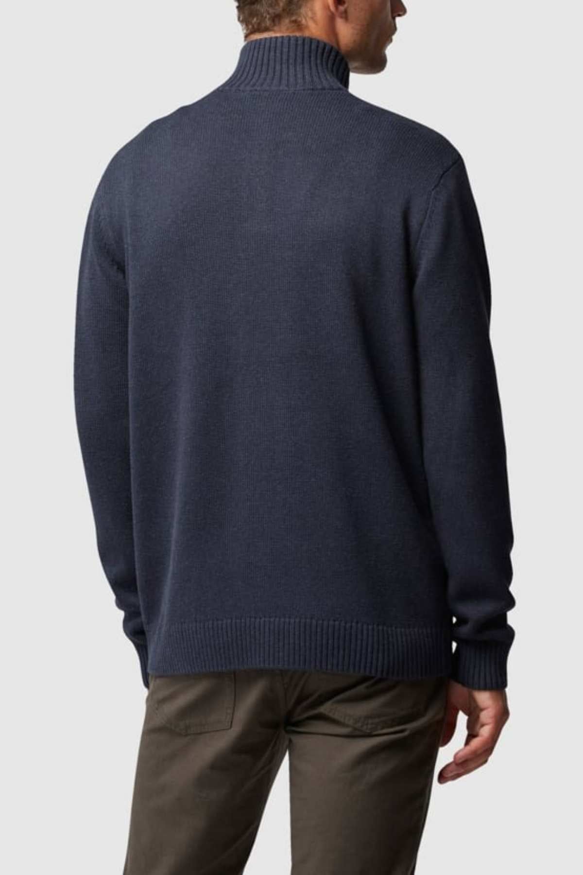 An individual with short, light hair is depicted from behind, wearing a dark blue long-sleeve Rodd & Gunn Merrick Bay Knit turtleneck sweater paired with olive green pants. The person is standing against a plain gray background.