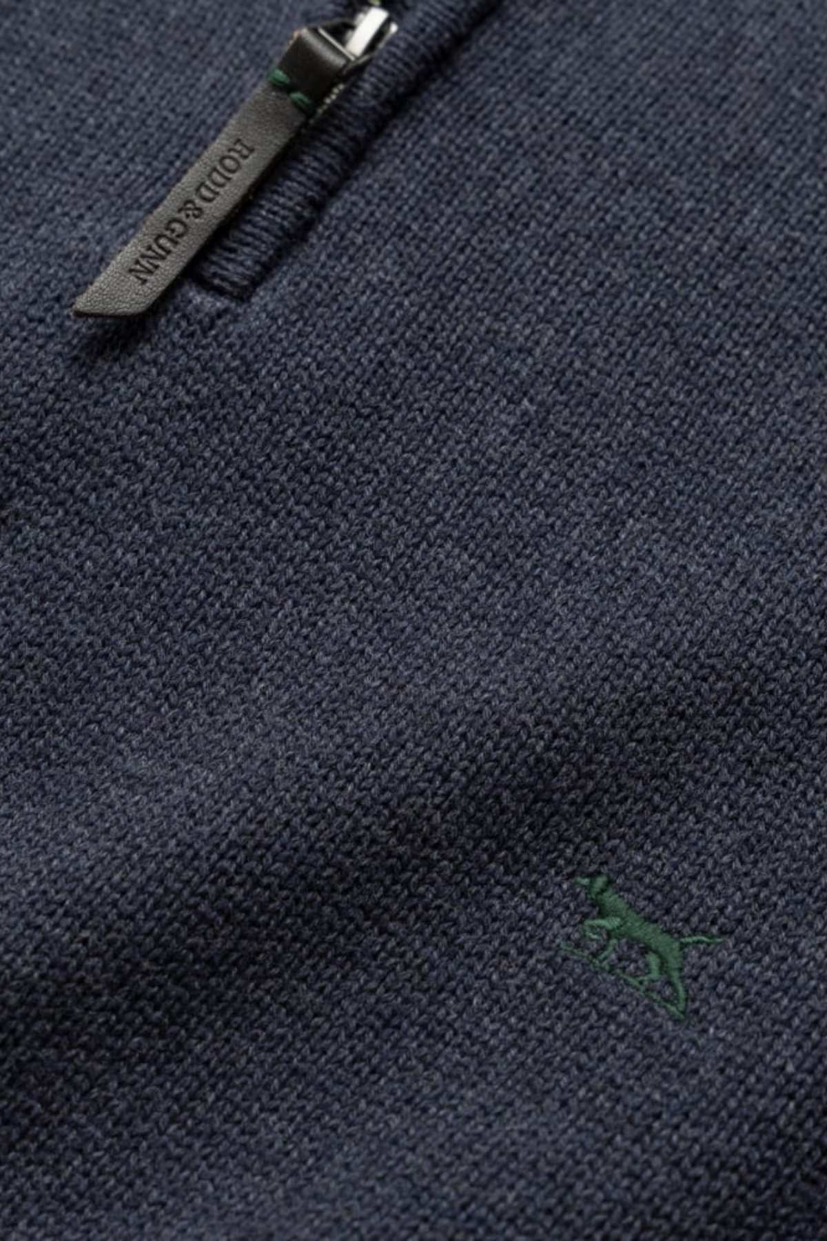 Close-up of a dark blue textured knit fabric featuring a partial premium YKK zip with a small green embroidered dog motif. The zipper pull is stamped with the text "BOBIDU CUENTIN," and the overall texture reflects the high quality characteristic of the Rodd & Gunn Merrick Bay Knit.
