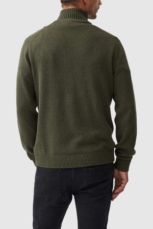 Rear view of a person wearing the Rodd & Gunn Merrick Bay Knit, a forest green high-neck sweater made from 100% cotton with ribbed cuffs and hem, paired with black jeans. The cozy jersey knit design contrasts against a light gray background.