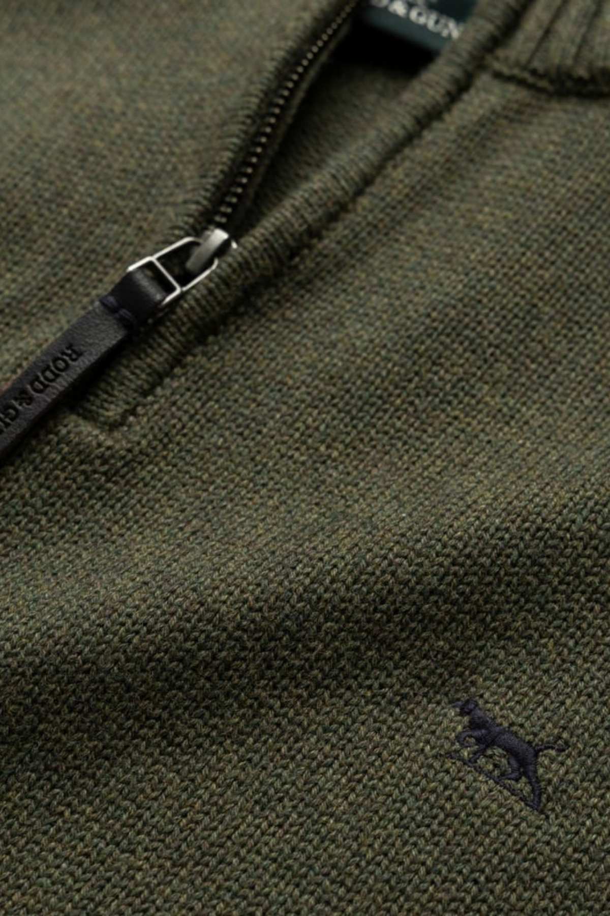 Detailed view of the Rodd & Gunn Merrick Bay Knit shows its textured olive green 100% cotton fabric, a premium black YKK half-zipper with visible stitching, and a small embroidered dog logo in dark color on the lower right.