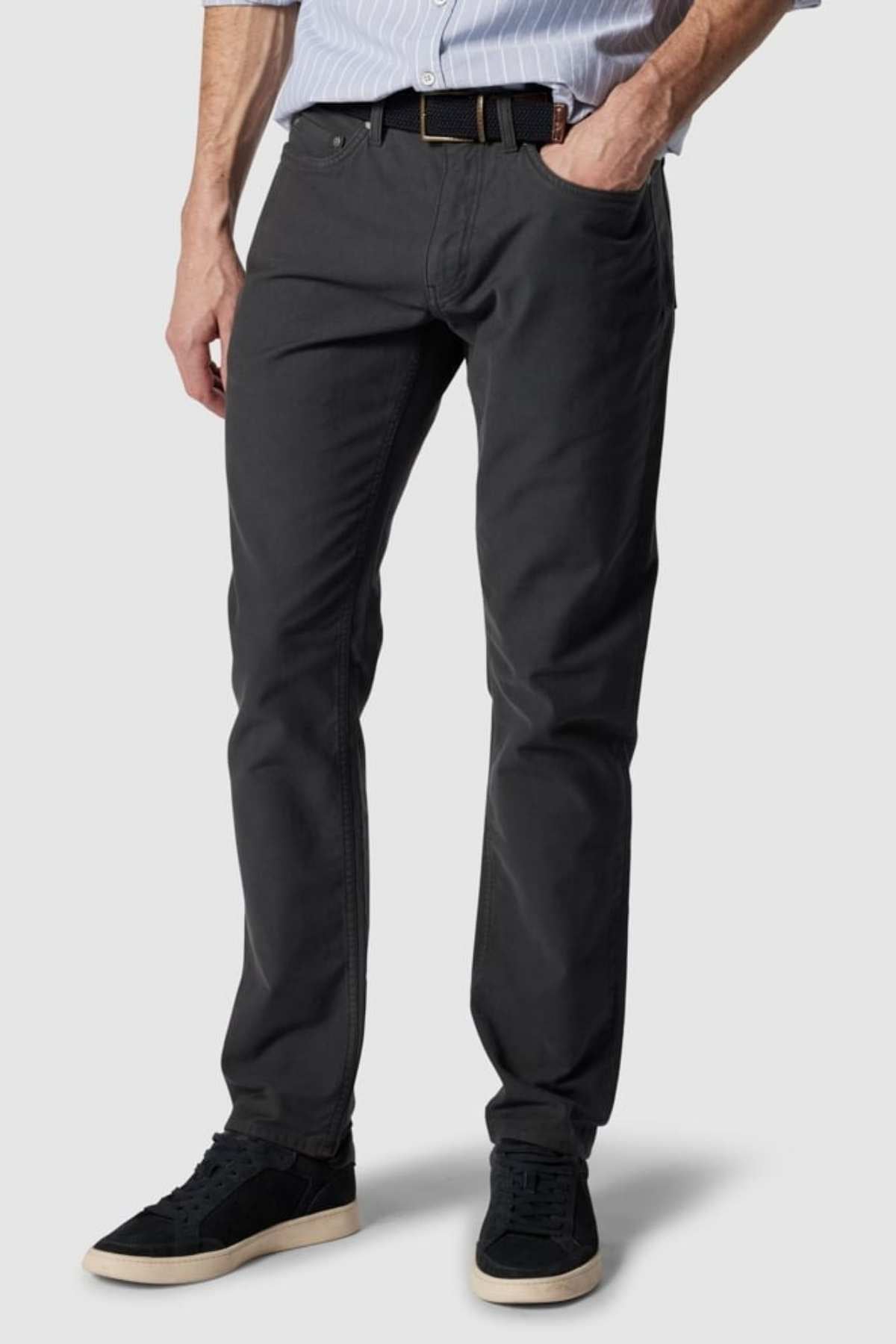 Wearing Rodd & Gunn Motion 2 Straight Jeans in dark gray with a YKK zip fly and comfort stretch, and complemented by a light blue striped shirt and black sneakers, the man stands coolly with his left hand in his pocket against a plain white background.