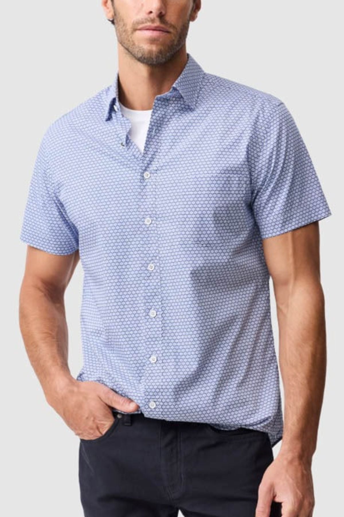 A man wears a Rodd & Gunn Blumine Sports Fit Shirt, featuring a light blue pattern and slim fit 100% cotton design, over a white undershirt with one hand in his dark pants pocket, against a plain light gray background.