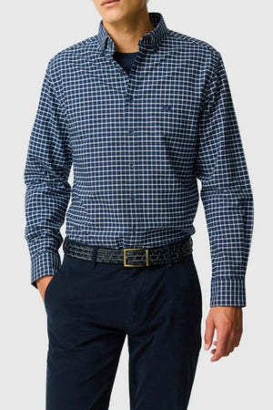 A man wears the Rodd & Gunn Gunn Check Oxford 3 Sports Fit Shirt, a long-sleeve blue and white check Australian cotton style, tucked into dark navy trousers with a black belt. The plain light gray background and his head are partially out of frame.