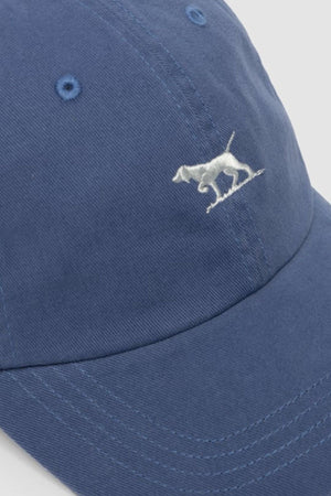 A close-up of the Rodd & Gunn Gunn Cap, a blue cotton twill cap with white stitching and a small embroidered pointer dog logo on the front.