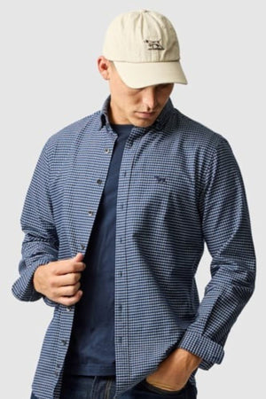 Wearing the Rodd & Gunn Gunn Check Oxford 5 Sports Fit Shirt in blue checks over a navy tee, jeans, and a cream baseball cap, a person looks down adjusting their shirt against a plain light gray background.
