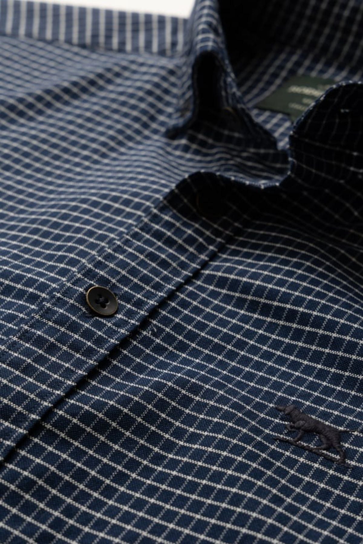 A close-up of the Rodd & Gunn Gunn Check Oxford 5 Sports Fit Shirt shows blue checkered fabric with white lines, dark buttons, and a small embroidered animal logo, crafted from premium Italian fabric for a tailored sports fit.