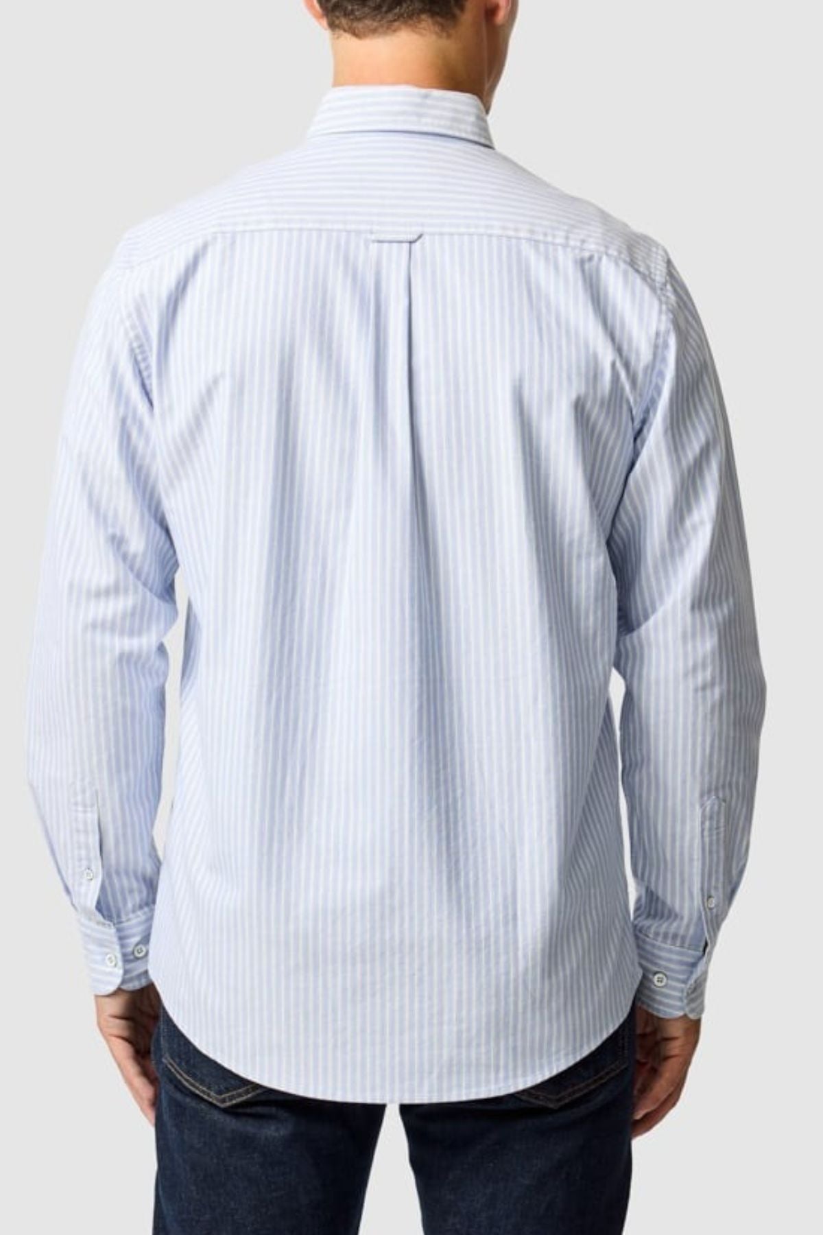 A man, seen from the back against a plain light gray background, wears the Rodd & Gunn Gunn Oxford Stripe 4 Of Shirt in light blue and white vertical stripes with dark jeans.