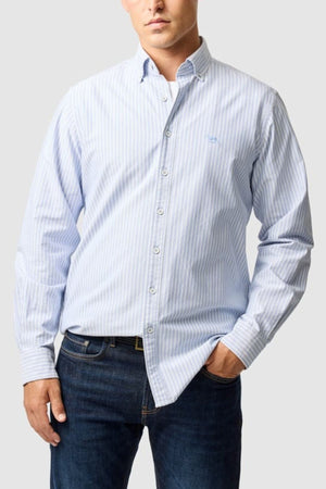 A man wears the Rodd & Gunn Gunn Oxford Stripe 4 Of Shirt in light blue and white pre-washed cotton with long sleeves and dark blue jeans, standing with one hand in his pocket against a plain light gray background.