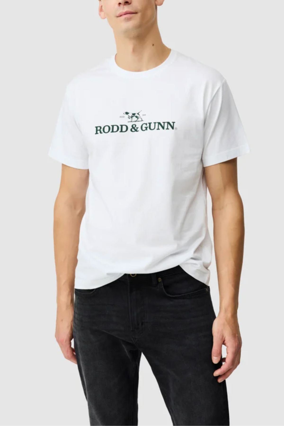 A man wearing the Rodd & Gunn The Logo Tee 2.0 and black pants stands against a plain light gray background, his head partially out of frame to highlight this classic menβs sports t-shirt style.