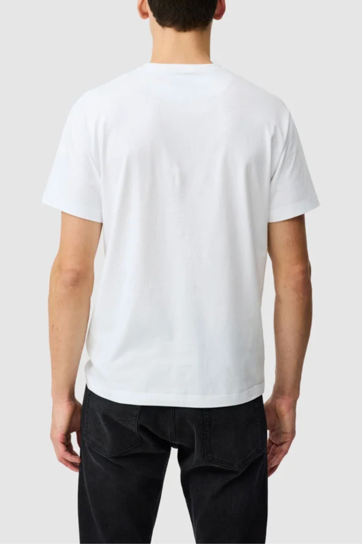 A person stands with their back to the camera in a plain white Rodd & Gunn The Logo Tee 2.0 and black jeans, set against a light gray background. This men's tee is made from 100% cotton for all-day comfort.