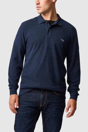 A man wearing a Rodd & Gunn Long Sleeve Sports Fit Gunn Polo in navy, made from breathable pique mesh fabric, and dark blue jeans, stands against a light gray background.